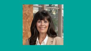 Headshot bordered in teal featuring a woman with long dark hair beige blazer and white blouse standing in front of lobby windows with brick accents