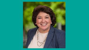 Headshot of Latina woman with brown curly short hair, grayish blue blazer and cream blouse in front of greenery and teal border