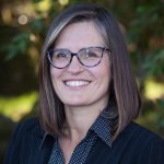 Headshot of white woman with salt and pepper shoulder length hair glasses navy and white polka dot blouse and pinstripe navy jacket