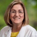 Headshot of white woman with brownish blonde hair glasses and yellow blouse underneath a white lab coat with greenery in the background