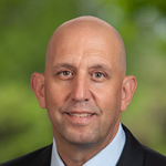 Headshot of white male with suit and tie and greenery in the background