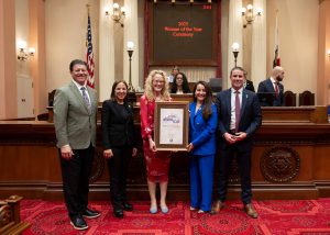 Four people standing in a line, from left to right, tall man brown hair in suit, woman in black suit, woman with blonde hair in red dress holding a sign, woman in blue suit holding award, man in blue suit arms crossed in front of him