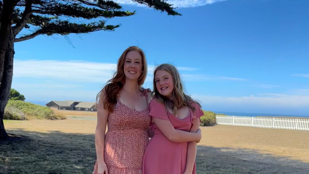 Mother and daughter wearing peachy pink sundresses pose under tree with coast line in the distance