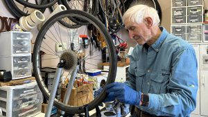 White man with white hair works on the wheel of a bicycle inside a workshop