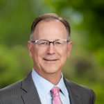 White man with glasses suit and tie in front of a greenery backdrop