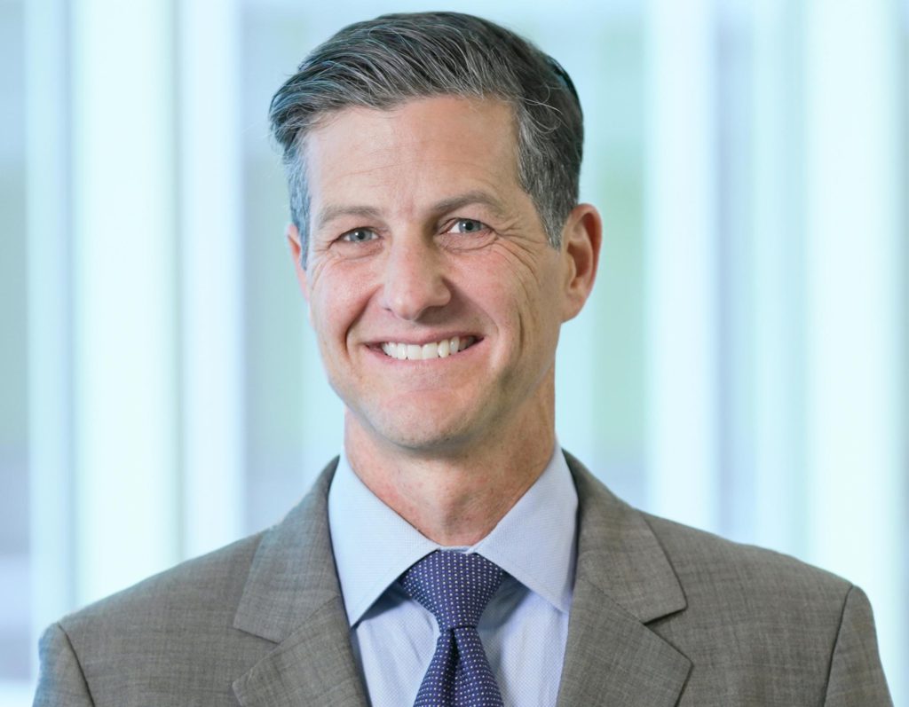 Doctor headshot with salt and pepper hair, wearing grey blazer, blue button up, and dark blue tie