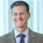 Doctor headshot with salt and pepper hair, wearing grey blazer, blue button up, and dark blue tie
