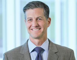 Doctor headshot with salt and pepper hair, wearing grey blazer, blue button up, and dark blue tie