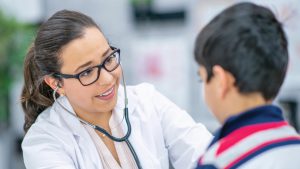 Latina physician with stethoscope and eyeglasses examines Latino boy wearing red, white and blue-striped inside exam room