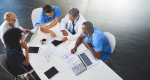 Team of medical workers sitting and meeting with laptops around table. Doctors and staff discussing papers and test results. Healthcare experts handling daily tasks and duties.
