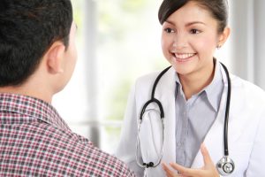 Latina doctor in white coat and stethoscope smiles at male patient with checkered shirt whose back is to the camera