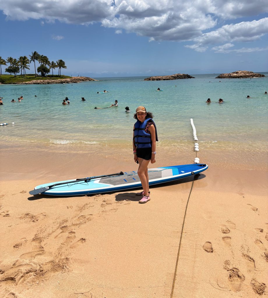 A teenage girl stands next to a paddleboard at the beach