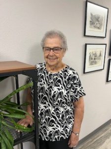 Senior Caucasian woman wearing black glasses. She has short grey hair and is wearing a black and white short-sleeve top.