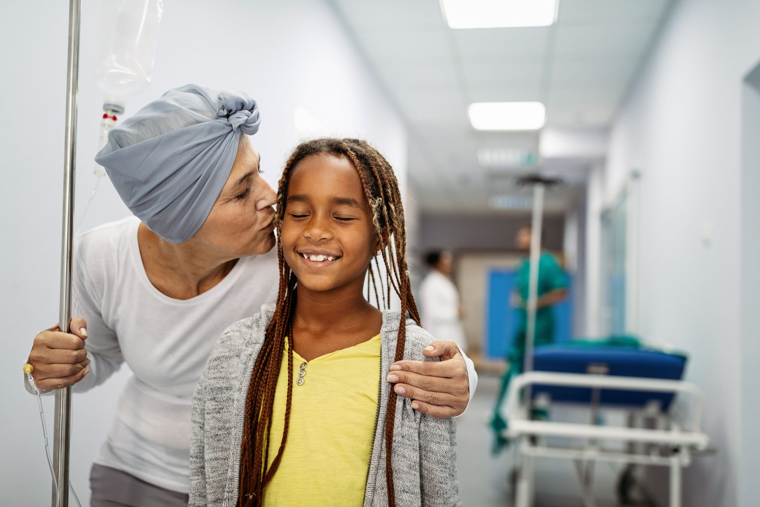 Becker’s Oncology_2025 Woman holding IV stand, wearing headwrap kissing young girl who is smiling