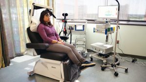 A woman sits in a large room with lots of natural light sitting in a exam chair while connected to several machines