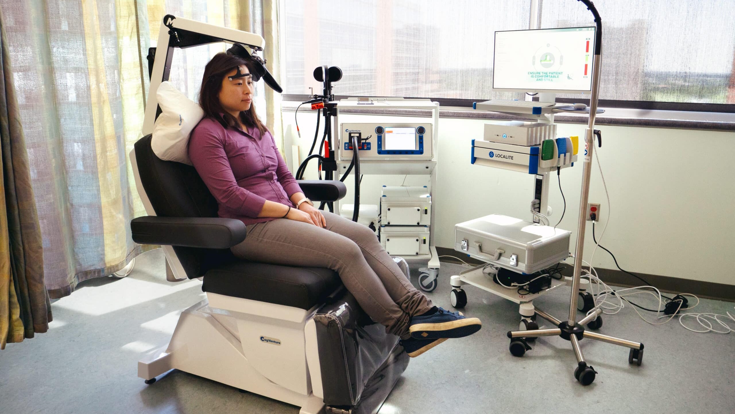 A woman sits in a large room with lots of natural light sitting in a exam chair while connected to several machines