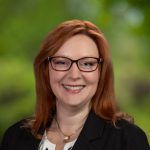 young woman with red hair smiling with glasses and green back drop
