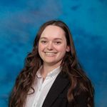 photo of smiling young woman, brown hair, with blue background, black suit 
