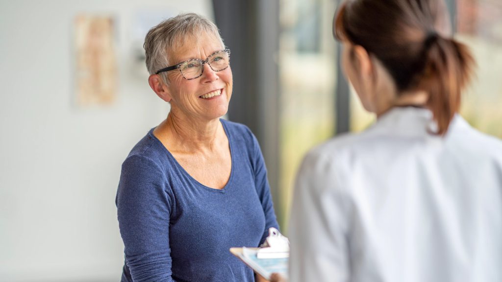 Older woman with short slat-and-pepper hair and blue V-neck long sleeve T-shirt consults with physician with reddish hair in a ponytail and a white lab coat