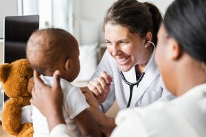 Woman doctor with dark-haired ponytail, white lab coat and stethoscope examines baby with mother and teddy bear on either side of baby