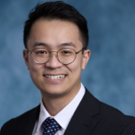 Asian man, smiling, with glasses blue background wearing a black suit