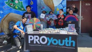 Teenage children holding decorative fans pose behind a table and in front of a multicolored mural on a city sidewalk