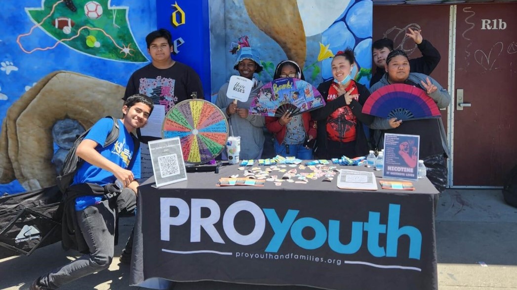 PRO Youth and Families Group Teenage children holding decorative fans pose behind a table and in front of a multicolored mural on a city sidewalk