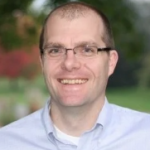 Man smiling with shaved head, glasses, blue button up