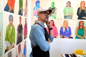A man with glasses and a driving cap stands reflectively in an art studio with colorful portraits on the walls