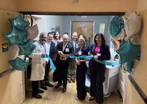 Business men and women and clinical team members surround a ribbon and a piece of medical equipment
