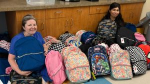 Two women sitting on the floor and leaning up against cabinets with about a dozen backpacks displayed in front of them