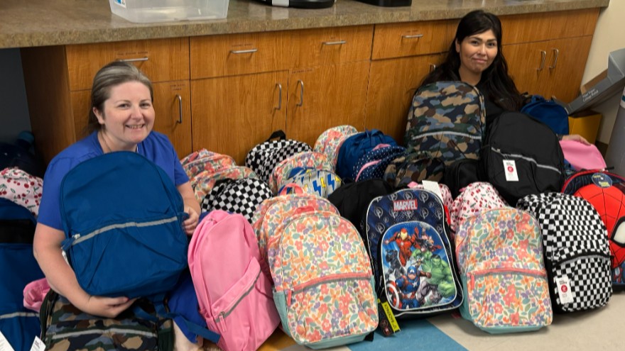 Two women sitting on the floor and leaning up against cabinets with about a dozen backpacks displayed in front of them