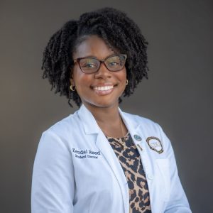Woman with dark hair and glasses, patterned blouse, and white coat 