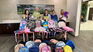 A group of women smiling in a lobby standing behind a table full of backpacks and school supplies