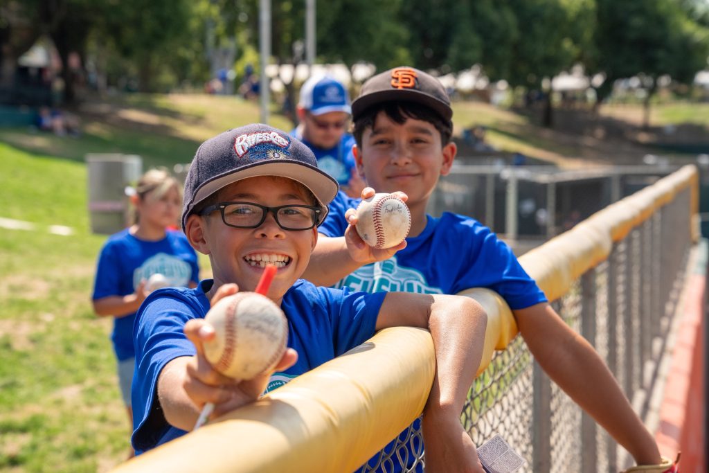 Two grinning boys with baseball caps and blue T-shirts holding baseballs lean on a fence