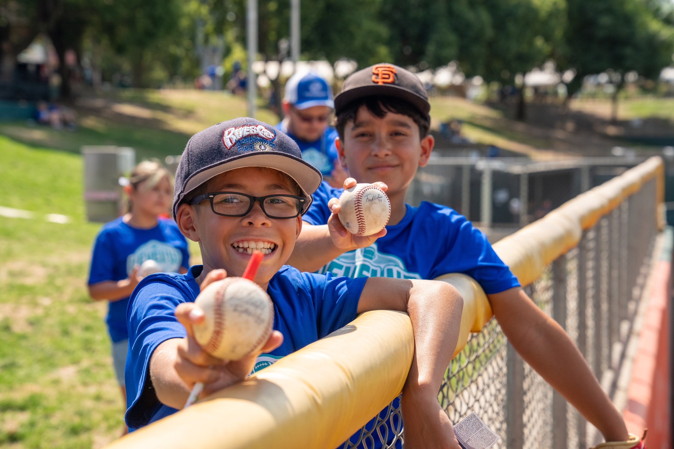 Two grinning boys with baseball caps and blue T-shirts holding baseballs lean on a fence