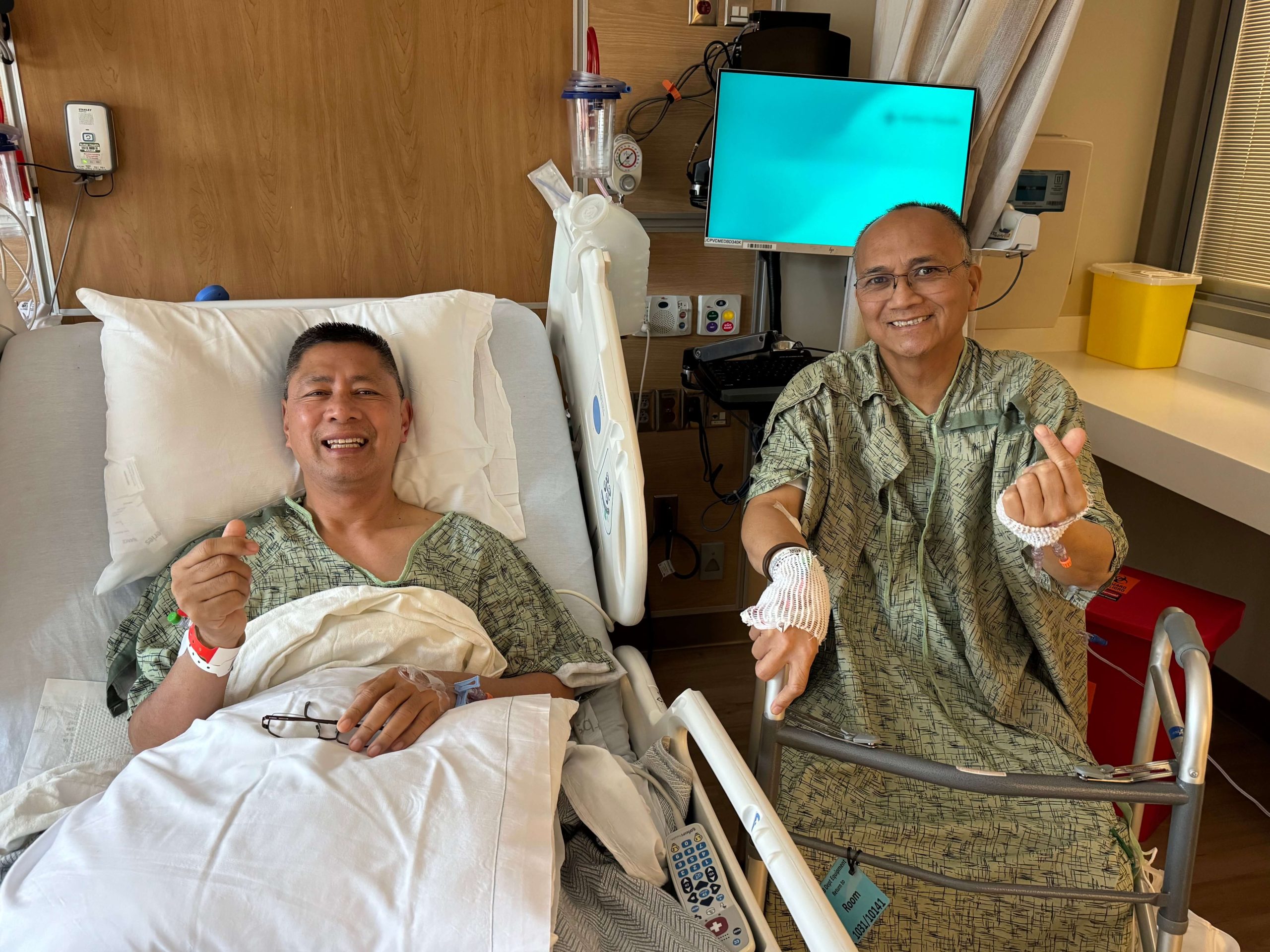 Two men in hospital gowns sit side by side in a hospital room. They are smiling