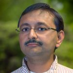Southeastern Asian man with glasses and moustache and red vertical striped shirt stands in front of greenery