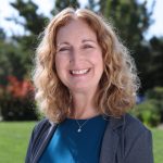 Woman with blonde wavy shoulder-length hair, blue corduroy blazer, blue blouse and silver pendant necklace stands with greenery in the background