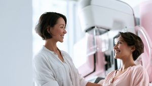 Ultra-realistic wide banner photograph of a female radiologic technologist calmly preparing a smiling woman patient beside a modern digital mammography unit, positioned on the far right third of the frame, bright clean imaging suite, soft pink accents, soft diffused window light, large clean empty negative space on the left side for text overlay, shallow depth of field, authentic clinical atmosphere